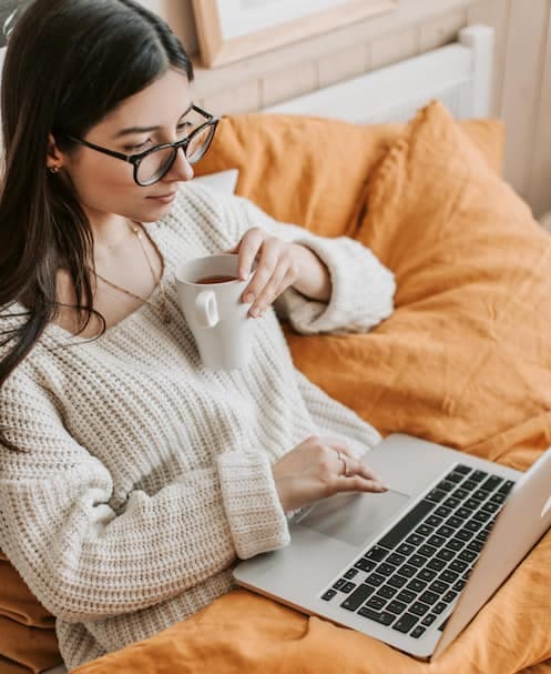 Relaxed woman sitting on bed with laptop, sipping coffee